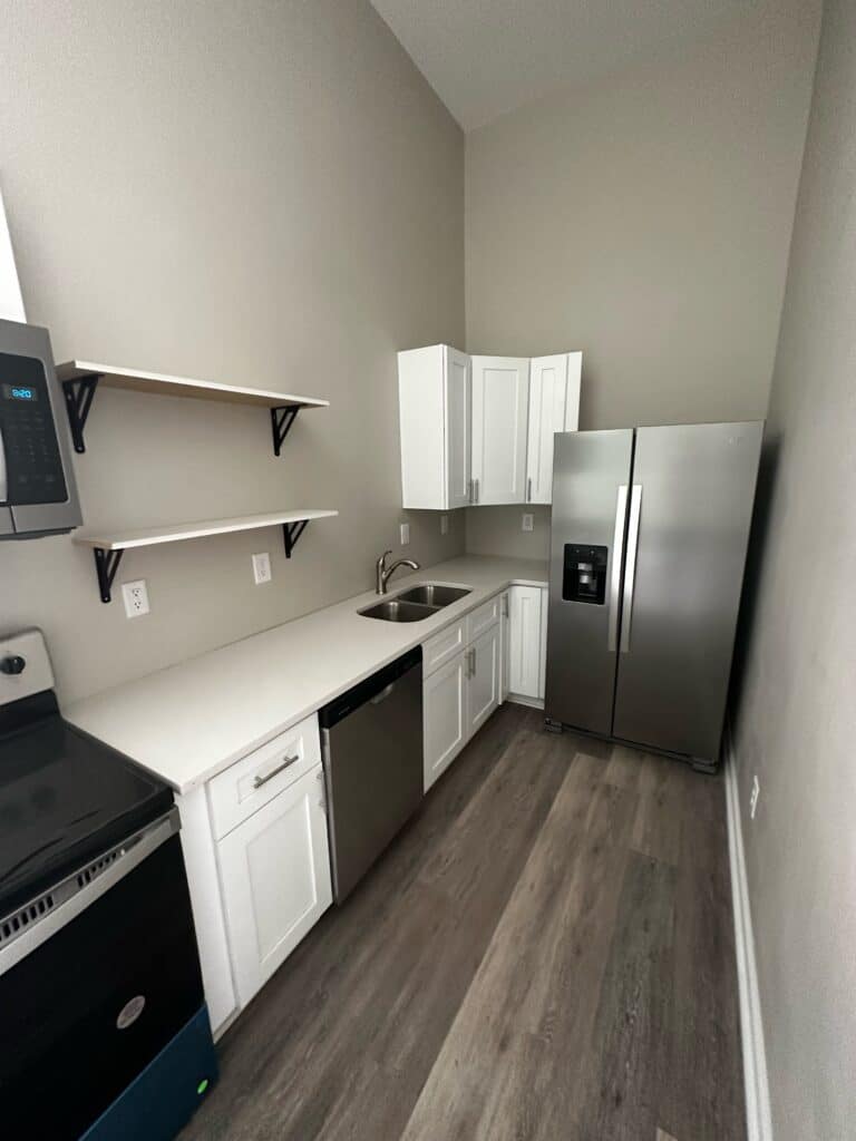 Modern galley kitchen with white cabinets, stainless steel appliances, open shelves, and wood-look flooring.