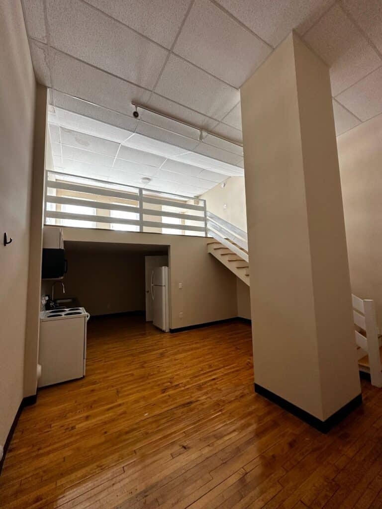 A small apartment with wood floors, a kitchenette, a white refrigerator, and a loft area accessible by stairs.