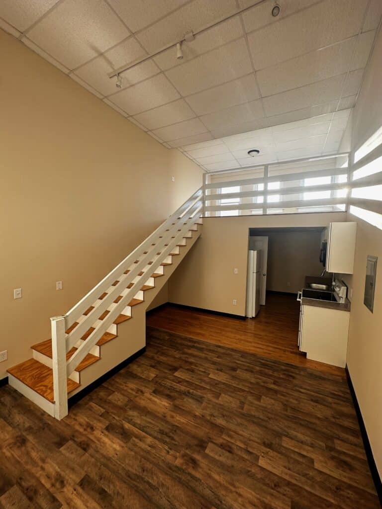 A modern loft apartment with wood flooring, a white staircase leading to an upper loft area, a small kitchen, and a refrigerator under a high ceiling.