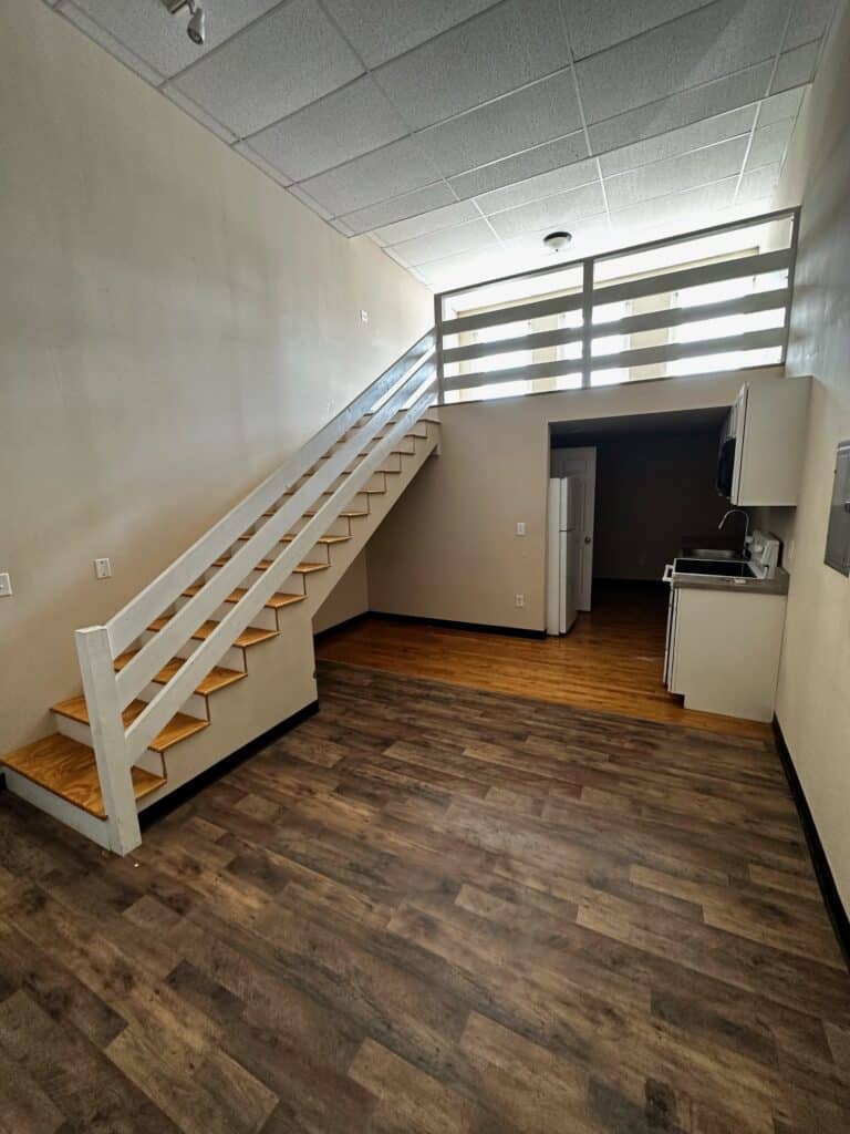 A small apartment interior with wooden stairs leading to a loft area, a kitchenette with a microwave and fridge below, and wood-style flooring throughout.