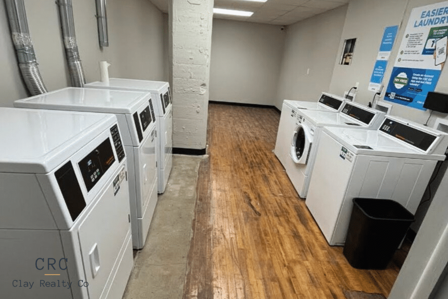 Laundry room with several white washing machines and dryers lined along the walls, wood-style flooring, and informational posters on the wall.