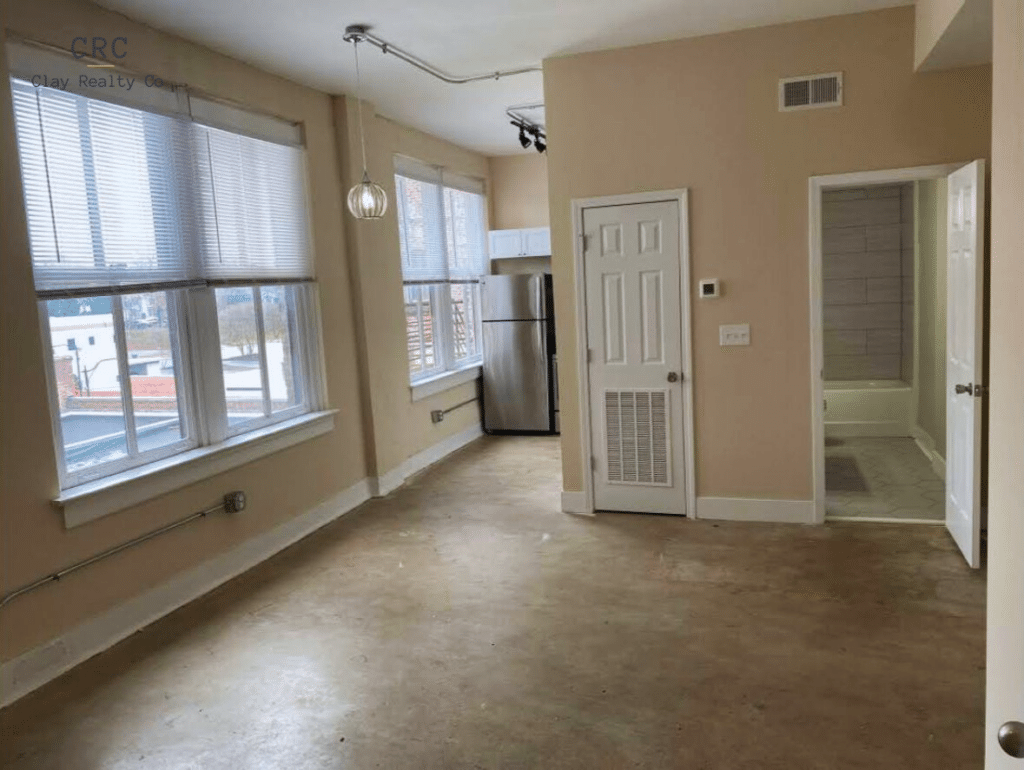 Empty room with beige walls and a concrete floor, featuring large windows, a small kitchen area with a stainless steel fridge, and an open door leading to a bathroom.