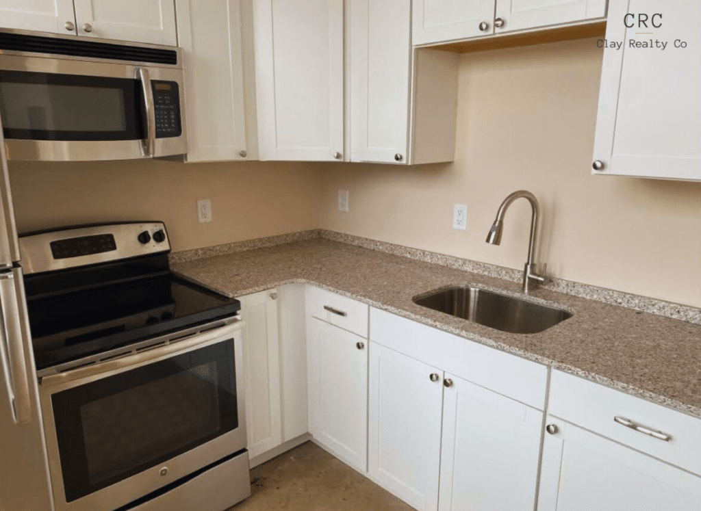A compact kitchen with white cabinets, a stainless steel oven, microwave, and a single basin sink under a granite countertop. Wall cabinets reach the ceiling.