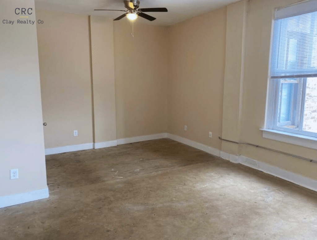 An empty room with beige walls and a cement floor, ceiling fan, and a window with blinds.