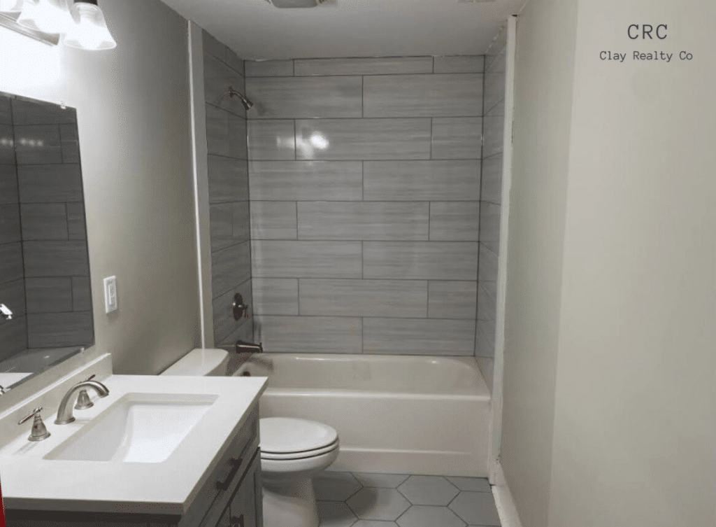 Modern bathroom with a gray tiled wall, a bathtub, a toilet, and a white sink with a countertop. Hexagonal floor tiles and a light fixture above the mirror.