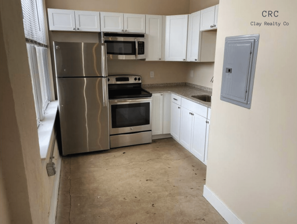 Small kitchen with white cabinets, stainless steel appliances, and beige walls. Includes a refrigerator, oven, microwave, and sink. Concrete floor and a window on the left.